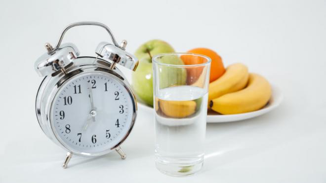 Reloj marcando las siete, vaso de agua y un plato de frutas al fondo (Freepik)