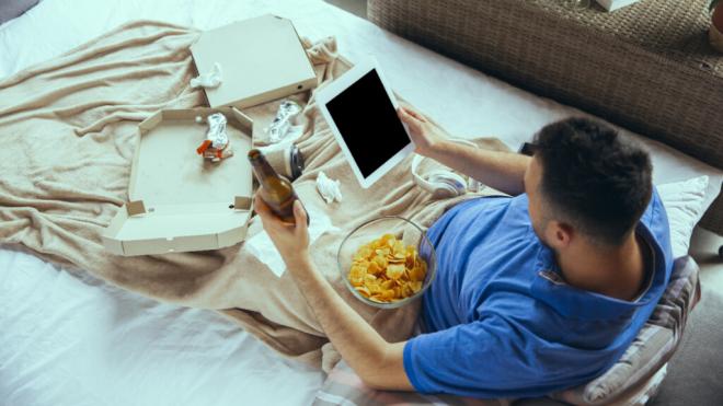 Hombre tumbado en la cama con restos de comida, con la tablet en una mano y una cerveza en la otra
