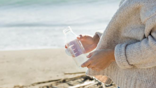 Mujer con una botella de plástico vacía en la playa (Foto: Freepik)