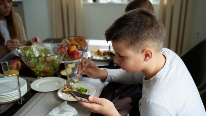 Niño comiendo en familia mientras está con el móvil (Fuente: Freepik)