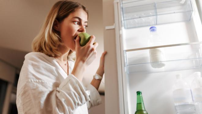 Mujer joven comiéndose una manzana mientras mira lo que hay en el frigorífico (Foto: Freepik)