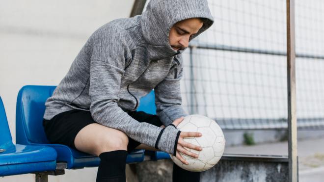 Hombre sentado en el banquillo de un campo de fútbol con un balón entre las manos (Foto: Freepik)