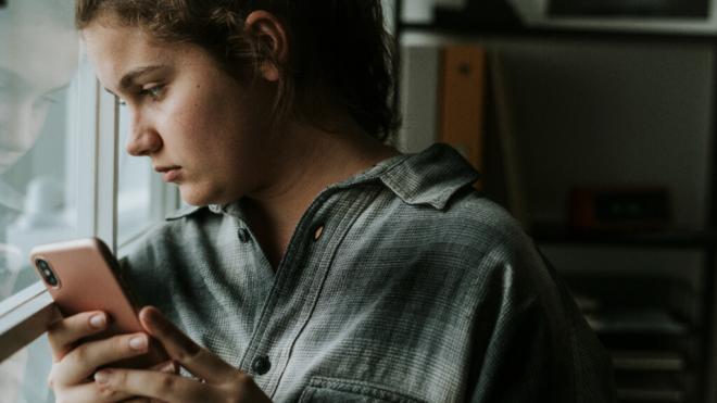 Mujer adolescente mirando por la ventana con un móvil entre sus manos (Foto: Freepik)