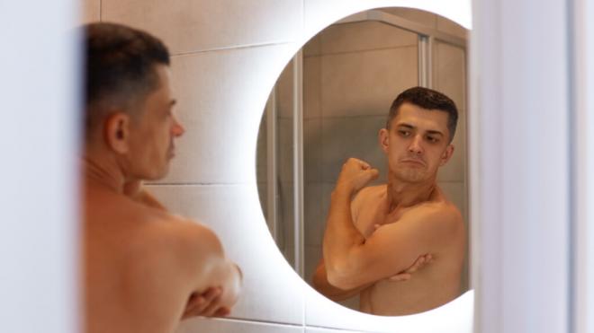 Hombre sin camiseta mirando en el espejo el grosor de su brazo (Foto: Freepik)