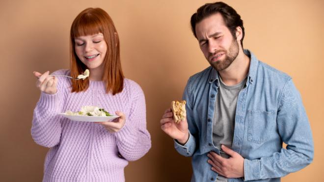 Mujer comiendo una ensalada feliz y hombre con una hamburguesa con malestar estomacal (Freepik)