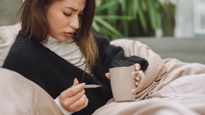 Mujer tumbada en el sofá sosteniendo un termómetro y una taza (Freepik)